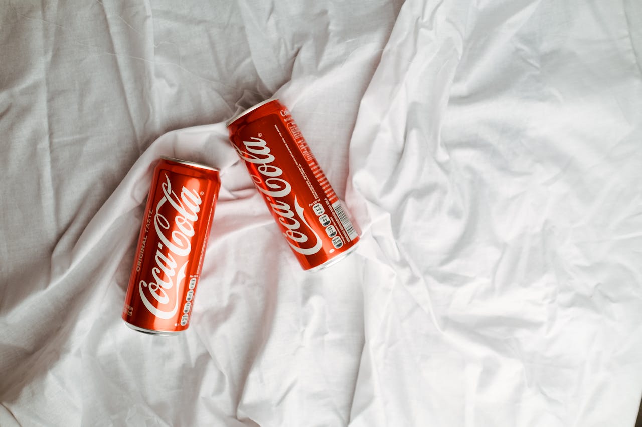 gallery-03 Two Coca Cola cans placed on a crumpled white textile, showcasing a classic beverage design.